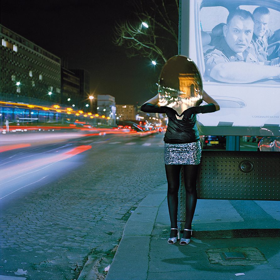 Sur les Champs-Elysées la nuit, au bord de la route et devant un panneau publicitaire, une femme en tenue de soirée tient un miroir ovale qui masque son visage et reflète à la place l'Arc de Triomphe.
