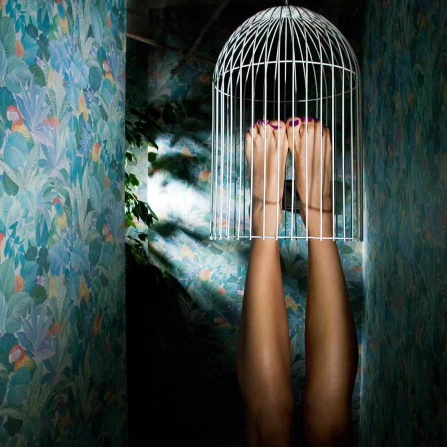 Dans un couloir au papier peint motif de la jungle, deux jambes feminines sont dans une cage à oiseaux.