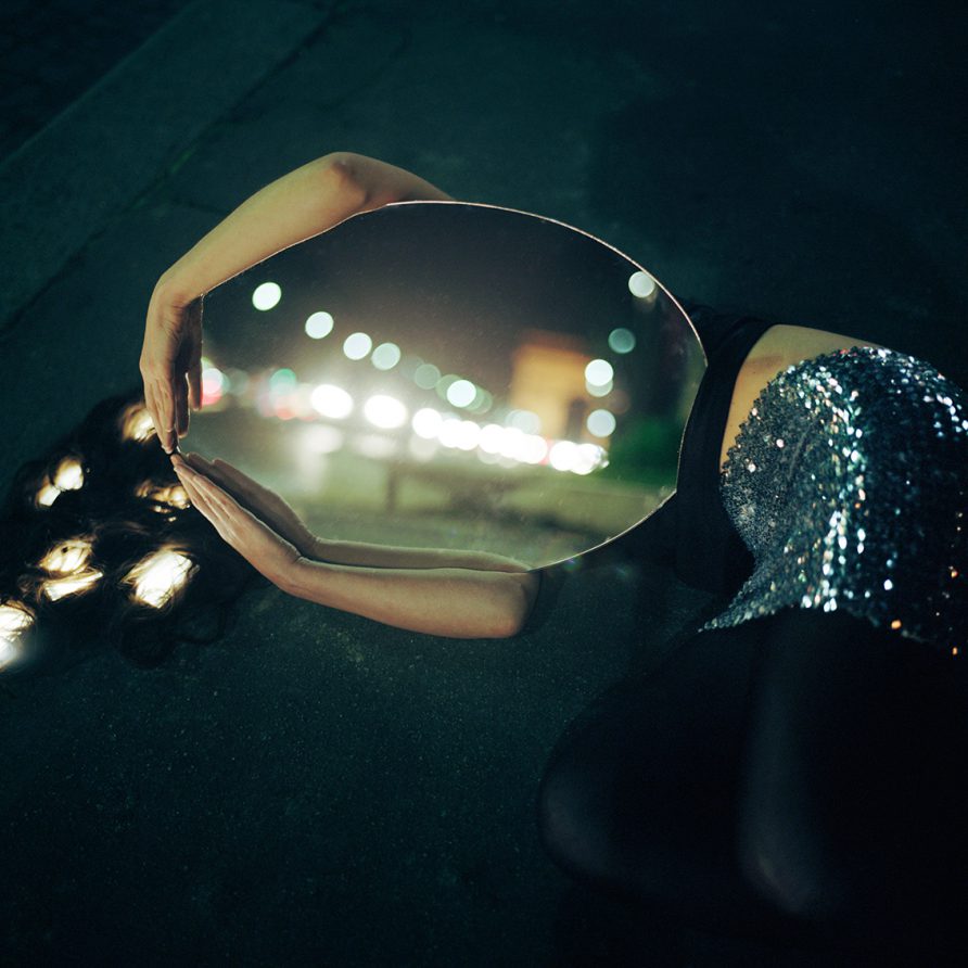 La nuit, une femme allongée au sol porte un grand miroir dans ses bras qui couvre son visage et reflète un flou de l'Arc de Triomphe.