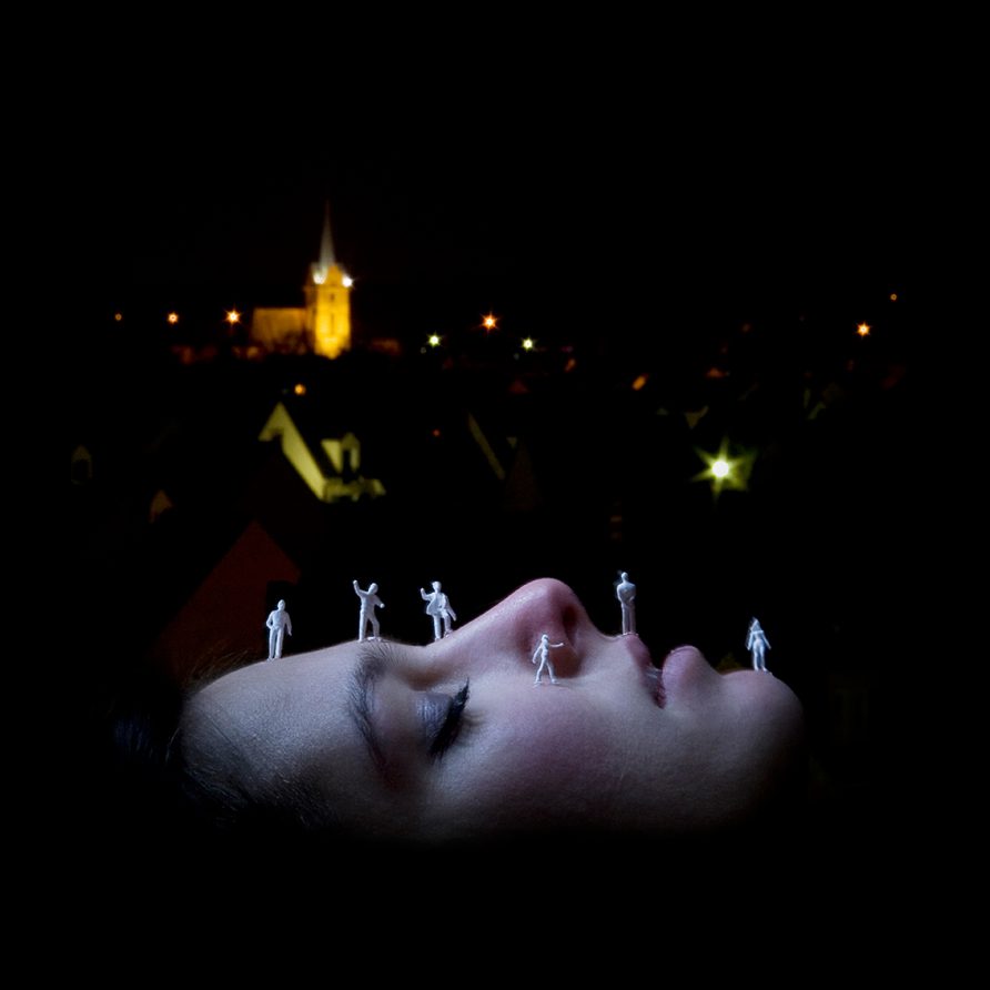 Le visage d'une jeune femme de profil sert de paysage sur lequel se promènent des personnages miniatures blancs. Depuis ce "relief féminin" on a une vue sur une petite ville plongée dans la nuit.