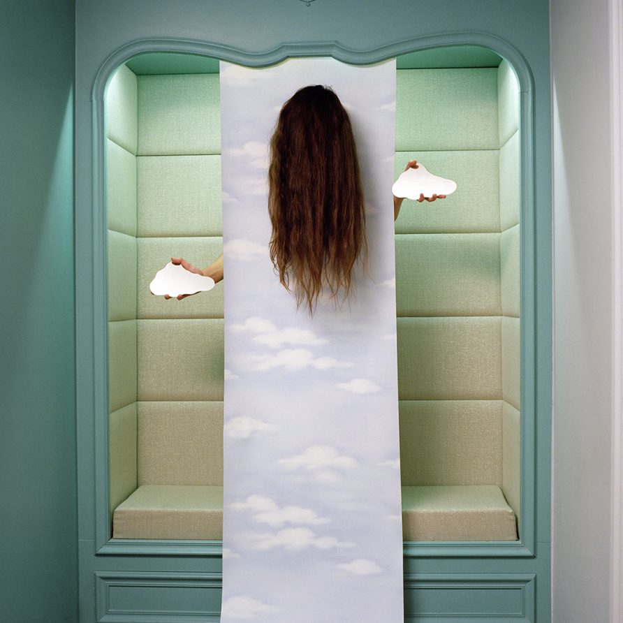 D'une alcôve part un papier-peint imprimé de nuages. De derrière ce papier sortent deux mains tenant des miroirs en forme de nuage ainsi qu' une tête dont on ne voit que la longue chevelure.