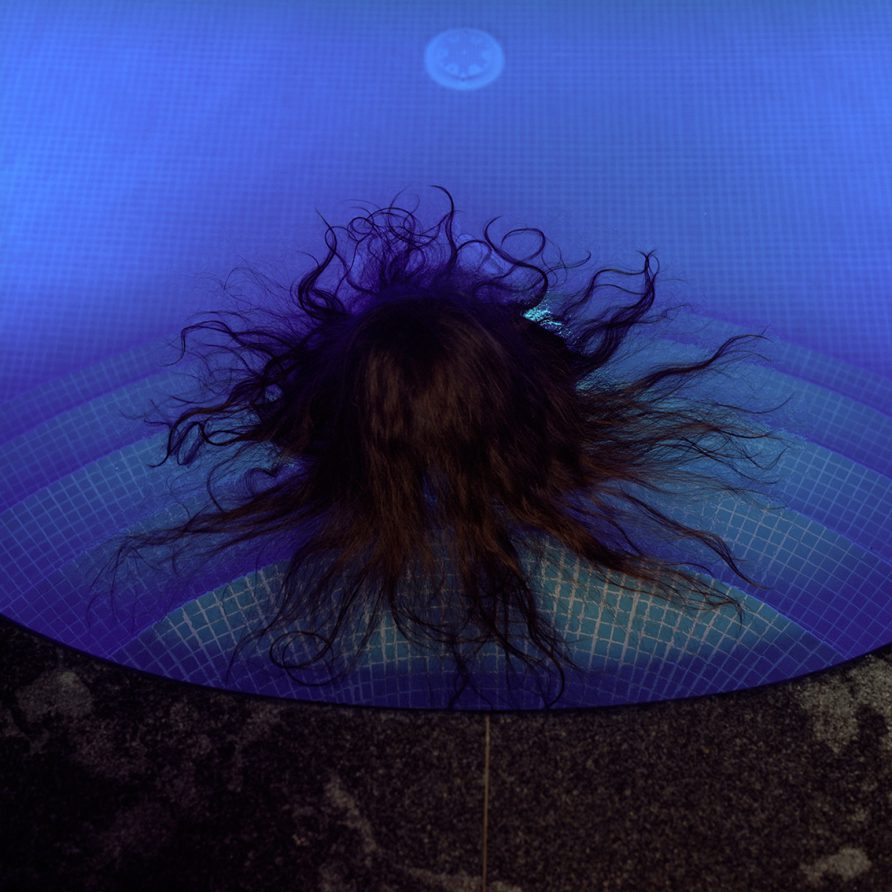 Dans l'eau d'une piscine, on ne voit du dessus qu'une tête brune dont les longs cheveux s'étalent en soleil à la suface de l'eau.