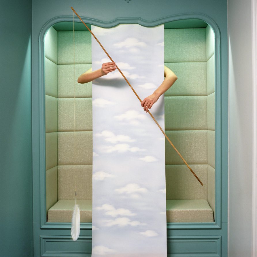 D'une alcôve part un papier-peint imprimé de nuages. Deux bras sortent de derrière ce papier et tiennent une canne à pêche au bout de laquelle pend une plume.