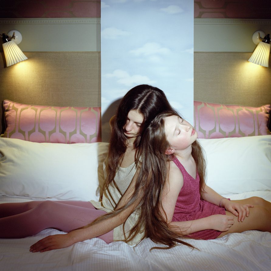 une fille et une jeune femme aux longues chevelures sommeillent dos à dos sur un lit. Derrière elles, un papier imprimé de nuages monte de leur lit.
