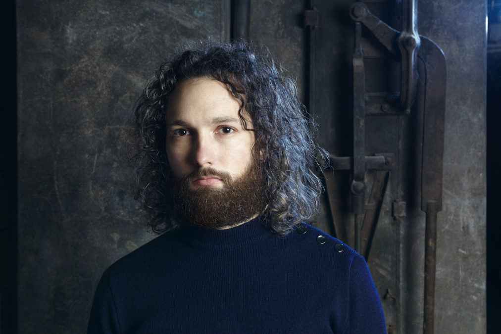 Portrait d'un homme barbu aux cheveux longs et bouclés dans une cave avec une lumière bleue.