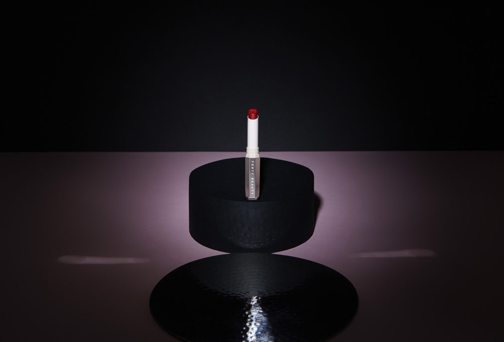 Nature morte en ambiance d'un rouge à lèvre Fenty Beauty ( = marque de la chanteuse Rihanna). Rouge à lèvre ouvert, raisin dévoilé, posé sur une estrade, tel une star. Jeux de lumière et miroir pouvant évoquer une boule à facettes...