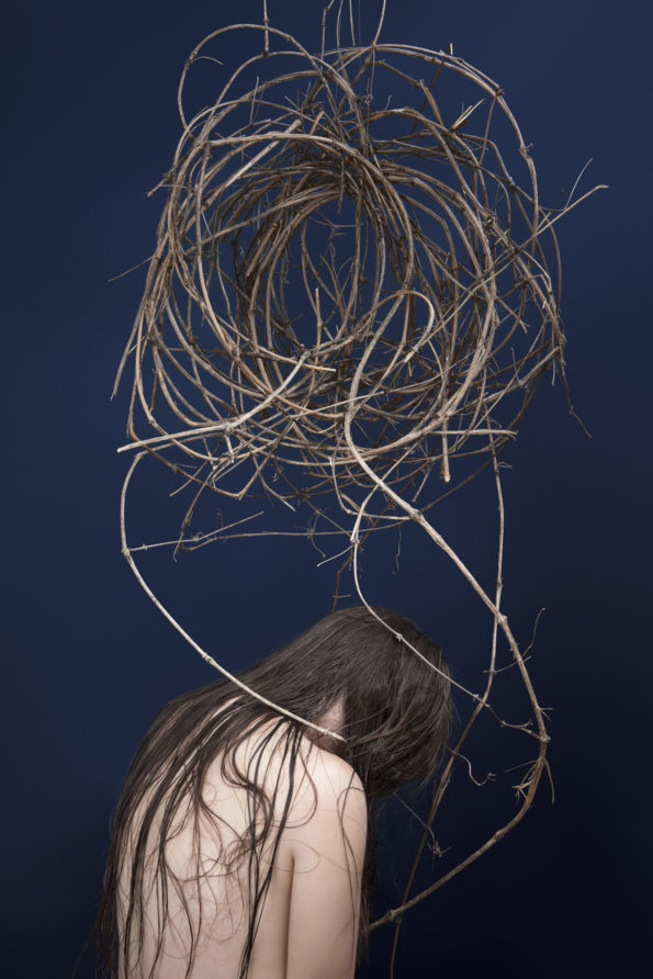 Femme aux cheveux longs et humides, positionnée de dos et courbée. Son visage est caché par ses cheveux et elle semble se ployer sous une boule de lianes formant un nuage au dessus de sa tête et efleurant son corps de certaines de ses tiges descendantes.
