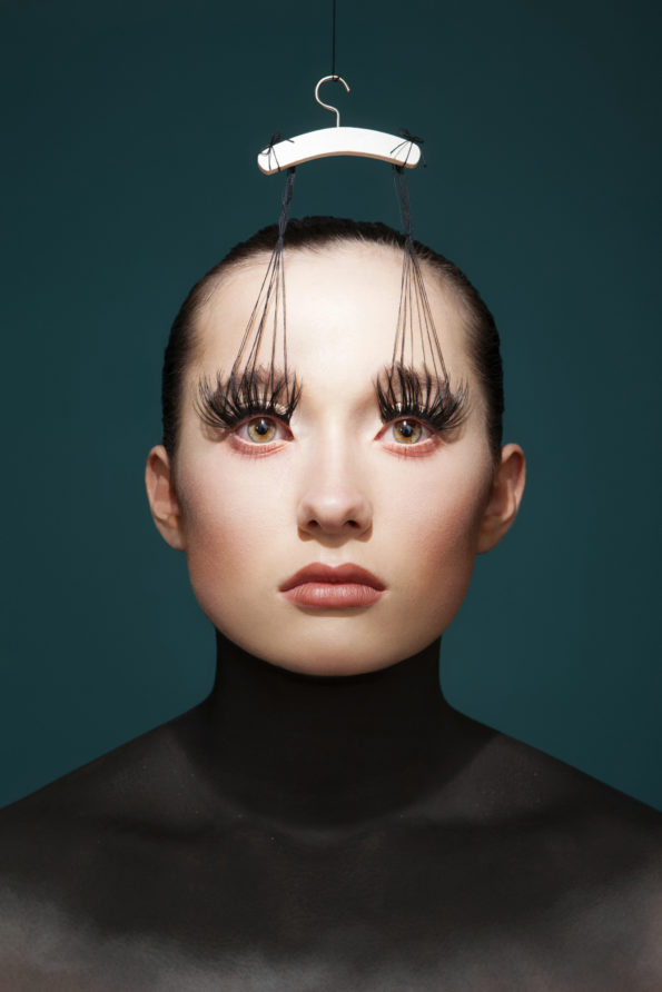 Image extraite de la série artistique : "Hypersensibilité".
Jeune femme les yeux grands ouverts, les cils prolongés en fils accrochés à un mini cintre. Cou sombre séparant ainsi la tête du reste du corps. fond bleu turquoise sombre.